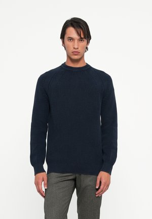 Suéter malha azul-marinho com textura canelada, gola redonda e mangas raglan longas, combinado com calças cinza bem ajustadas.