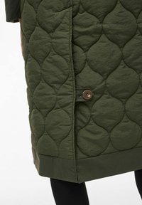 Olivenfarget kviltet jakke med teksturert mønster, rett snitt og knappdetalj på siden. Glatt stoff med matt overflate.