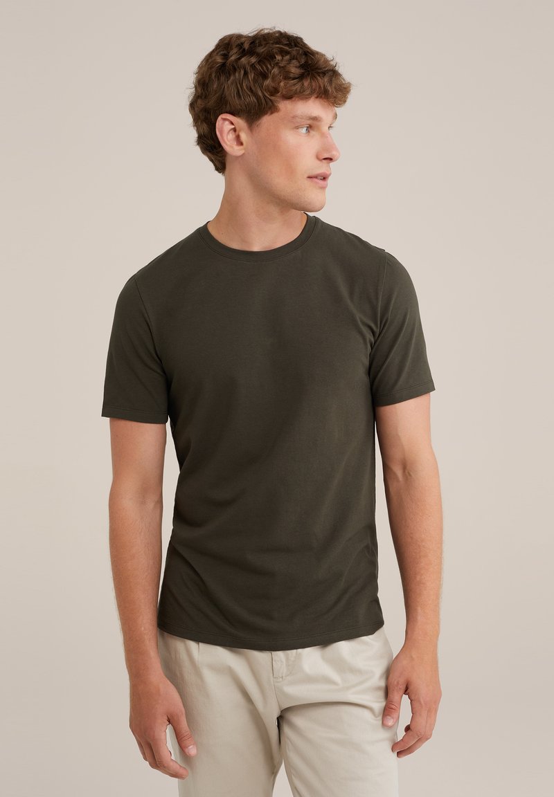 Camiseta de manga corta verde oscuro, con cuello redondo, hecha de algodón suave. El modelo la combina con pantalones de color claro, mostrando un ajuste casual.