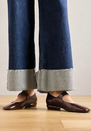 Brune lakk lær sko med utskjæringsdesign, lav hæl og ankelreim, kombinert med vide denimjeans med fransede kanter.