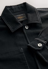 Chemise en denim noir avec un col structuré, des accents de coutures blanches, une fermeture à deux boutons, et un tissu texturé avec une étiquette.