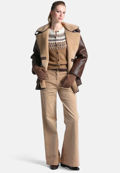 Femme portant une veste en cuir marron et en peau lainée, un cardigan à motifs, un pantalon beige en velours côtelé, des gants marron et des chaussures noires, debout les mains sur les hanches.