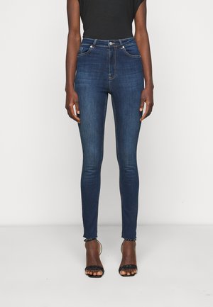 Person iført mørkeblå high-waist skinny jeans med rå kant kombineret med sorte flettede stroppesandaler med høje hæle og en sort top.