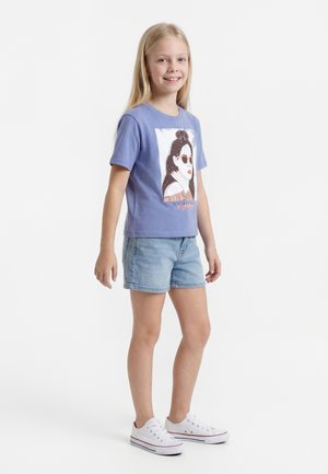 Smiješeća djevojka s dugom plavom kosom, nosi plavu majicu s printom ženskog lica, traper kratke hlače i bijele tenisice, stoji bočno.