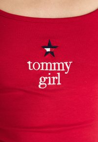 Červený bezrukávový top vyrobený z mäkkého materiálu, ktorý sa vyznačuje bielym vyšívaným textom "tommy girl" a čiernou hviezdou s červenými, bielymi a modrými akcentmi.