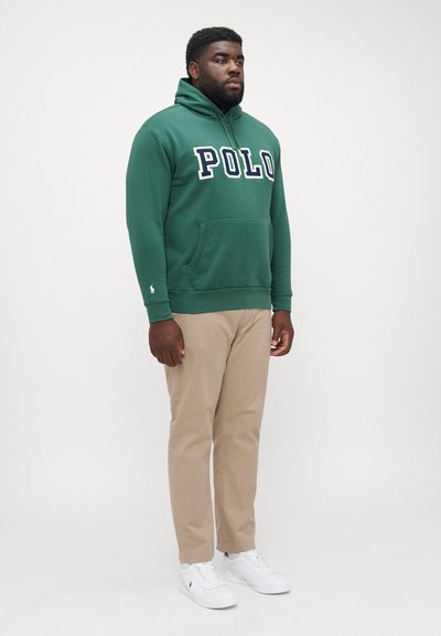 Felpa verde in cotone con logo "POLO" in lettere bianche, tasca frontale, polsini a costine, abbinata a pantaloni khaki e sneaker bianche.