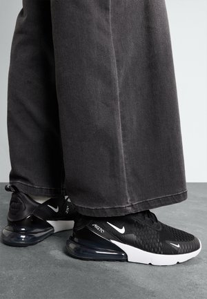 Osoba ubrana w czarne szerokie spodnie i czarne sneakersy Nike Air Max 270 stoi na szarej podłodze.