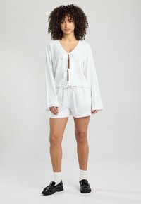 White linen top with ties at the neckline and long sleeves, paired with white shorts. The model wears black loafers and white socks.