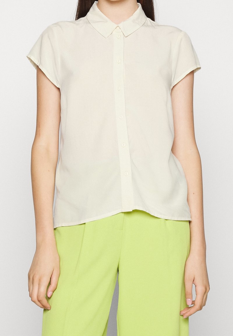 Femme portant une chemise beige à manches courtes boutonnée et un pantalon vert lime, debout contre un fond clair uni, les bras détendus.