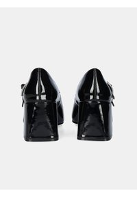 Zapatos de charol negro con tacón cuadrado, con una superficie lisa, que presentan un detalle de hebilla lateral y un diseño de punta ligeramente afilada.