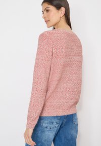 Femme aux longs cheveux bruns portant un pull rouge à taches blanches et un jean bleu, légèrement tournée sur le côté sur un fond blanc.