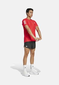 Camiseta atlética roja de manga corta con rayas blancas en los hombros, combinada con pantalones cortos negros y zapatillas deportivas blancas.