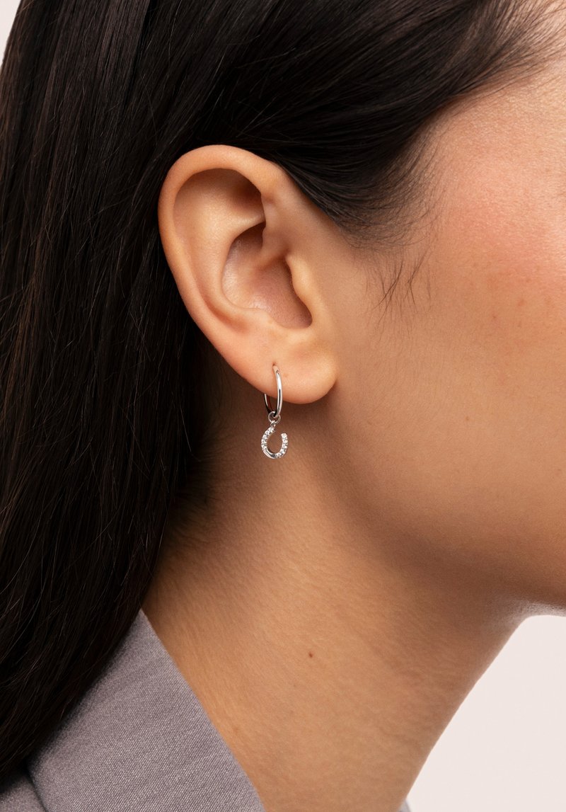 Silver hoopörhängen har en liten hängande berlock formad som en hästsko, dekorerad med klara stenar. Designen är elegant och minimal.