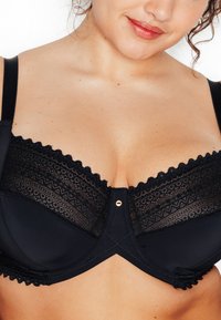 Reggiseno nero con parte superiore in pizzo a motivo a zigzag, bordi smerlati e un bottone decorativo dorato centrale. Spalline regolabili visibili.