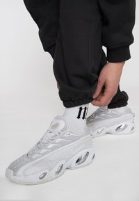 Biele tenisky Nike s textúrovaným sieťovaným zvrškom a priehľadnými vzduchovými vreckami. Čierne joggers s elastickými manžetami pri členkoch, nosené s bielymi ponožkami so značkovým logom.