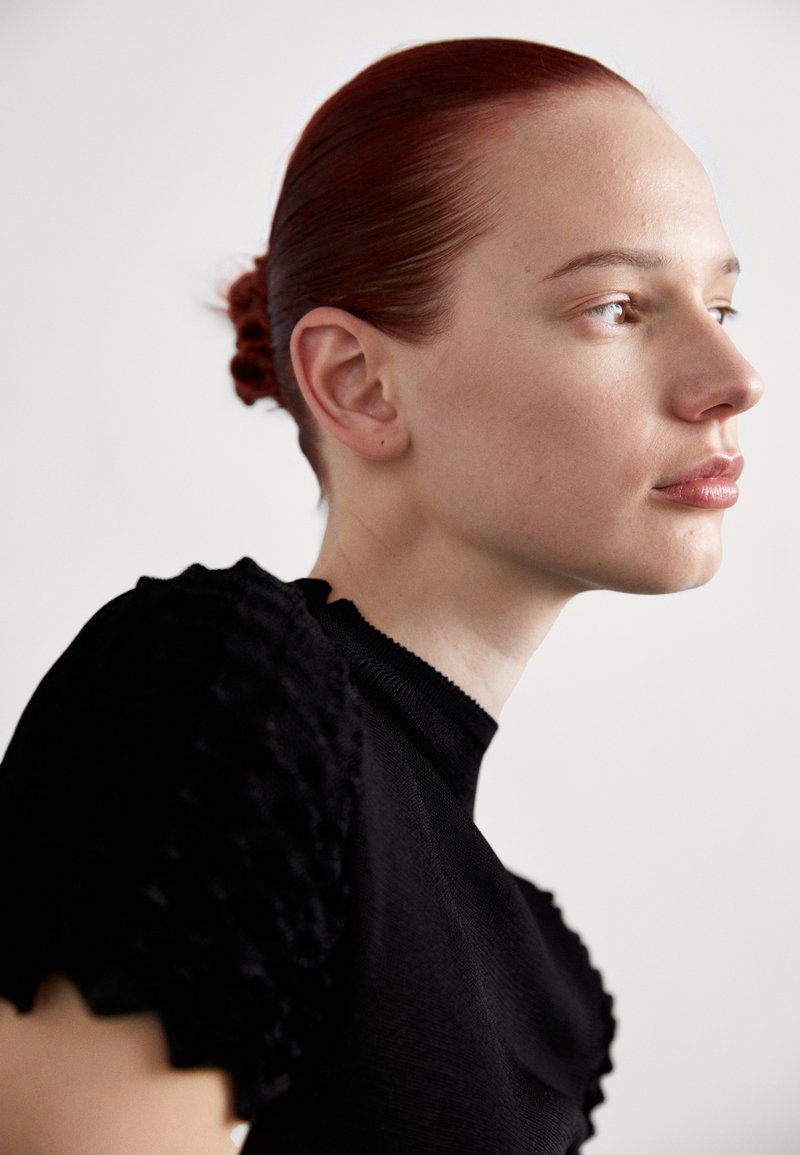 Top negro con ribetes y mangas con borde en forma de ondas, presenta un diseño ajustado. El modelo tiene el cabello rojo peinado hacia atrás y maquillaje minimalista, todo contra un fondo neutro.