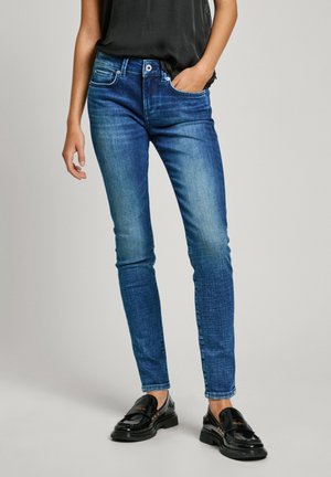 Person trägt blaue Skinny-Jeans, schwarze Loafer und ein schwarzes Hemd, steht mit einer Hand in der Tasche vor einfarbigem Hintergrund.