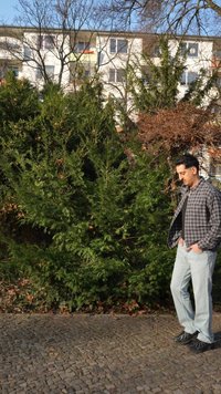 Hombre con camisa de cuadros y jeans de pie en un camino de adoquines junto a arbustos verdes, con edificios residenciales al fondo.