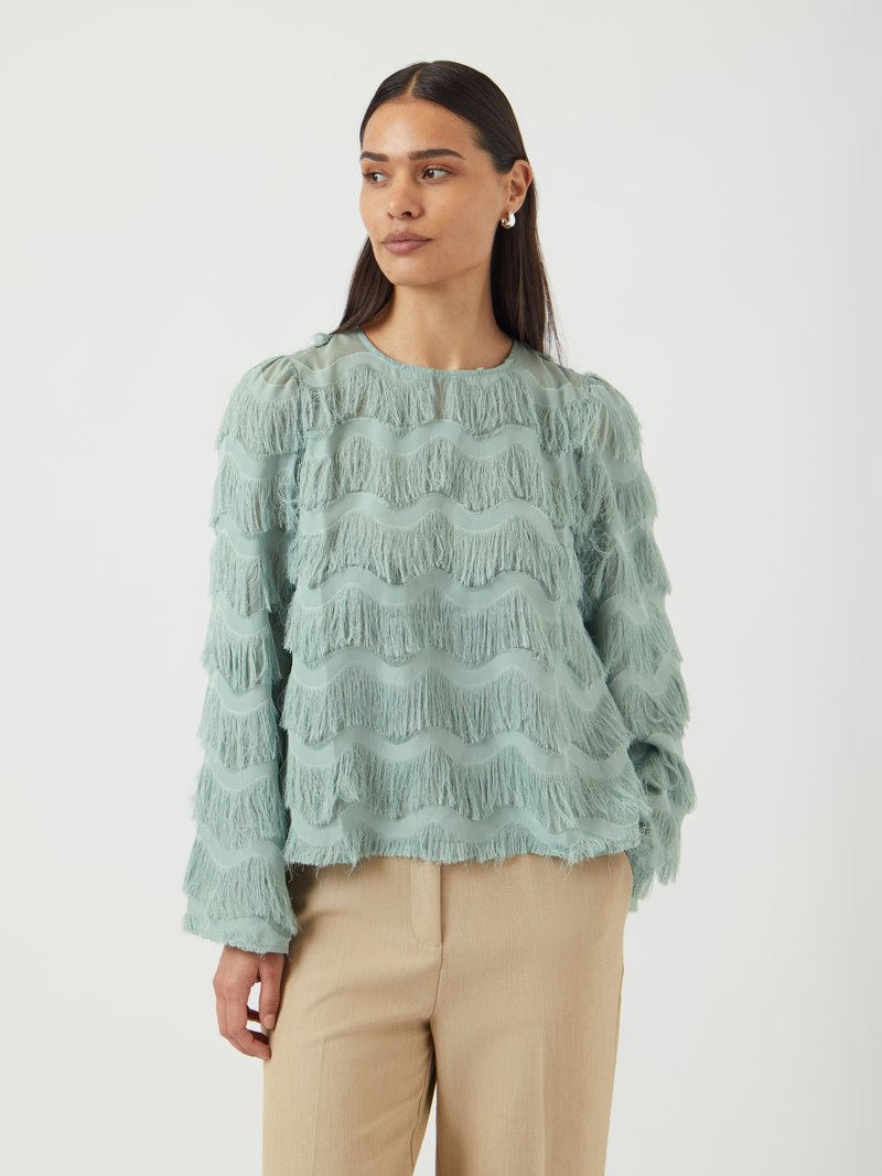 Top trasparente azzurro chiaro con dettagli frangiati ondulati; maniche lunghe e scollatura arrotondata; abbinato a pantaloni beige.