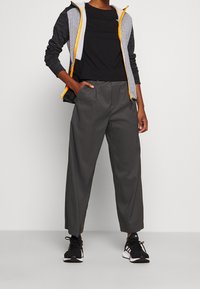 Persoon met een zwart shirt met lange mouwen, een wit vest met een patroon en een rits met oranje rand, grijze wijde broek en zwarte Adidas sneakers.