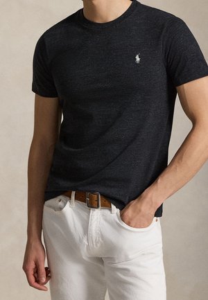 Mężczyzna ubrany w granatowy t-shirt z krótkim rękawem w ciemnym melanżu, ozdobiony małym, białym haftowanym logo, w połączeniu z białymi spodniami i brązowym paskiem.