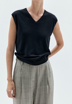 Person wearing a sleeveless black V-neck top tucked into high-waisted beige plaid pants with one hand behind their back.