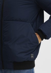 Marineblauwe pufferjack met een ritssluiting, ribgebreide manchetten, zijzakken en een logopatch aan de onderkant aan de linkerkant. Gladde textuur.