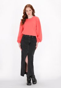 Top corallo a maniche lunghe con maniche a sbuffo abbinato a una gonna di jeans nera con spacco frontale. Modella che indossa stivali neri alla caviglia.