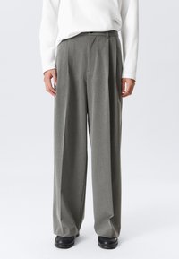 Personne portant un pantalon large à rayures grises, un haut à manches longues blanc et des chaussures noires, se tenant devant un fond uni.