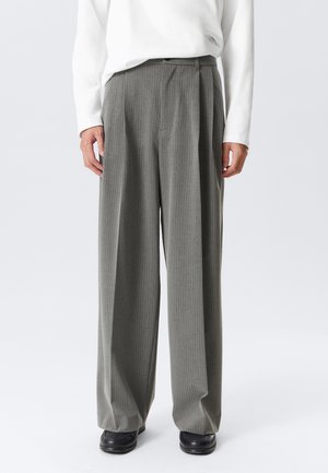 Person wearing gray pinstripe wide-leg trousers, white long-sleeve top, and black shoes, standing against a plain background.