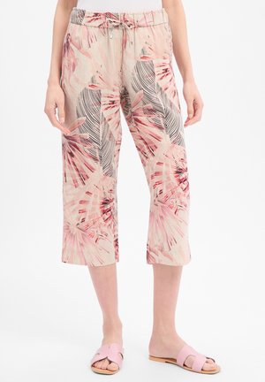 Woman wearing beige cropped pants with pink and black tropical leaf print and pink slip-on sandals, standing against white background.