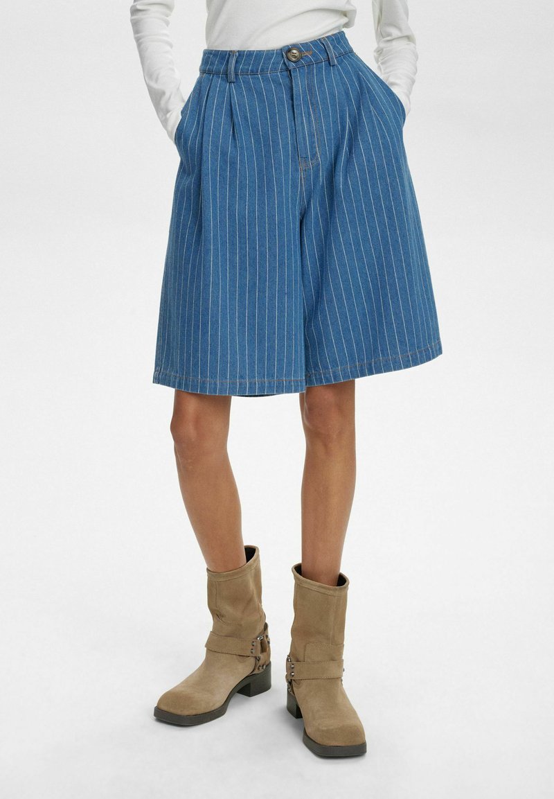 Kék farmer culotte, fehér függőleges csíkokkal, pliszírozással és gombos derékzárral. Bézs bokacsizmával párosítva.