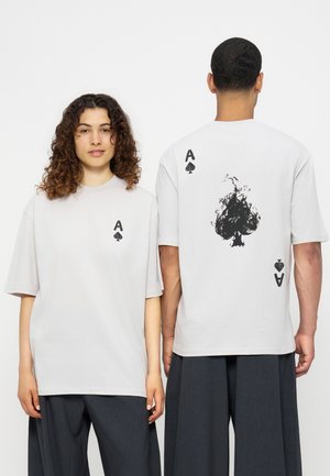 Dos personas llevan camisetas blancas con símbolos negros de as de picas, una mirando al frente con un símbolo pequeño, la otra de espaldas con una pica grande en llamas.