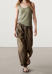 Olijfgroen geribbeld tanktop gecombineerd met losse, bruine broek met trekkoord. Model draagt zwarte sandaaltjes met bandjes; minimalistische achtergrond.