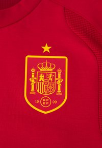 Κόκκινη αθλητική φανέλα με κίτρινο έμβλημα που διαθέτει περίπλοκα σχέδια, ένα στέμμα και ένα αστέρι από πάνω. Περιλαμβάνονται οι λέξεις "RF EF" και "1909" στο έμβλημα.