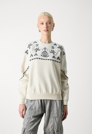 Cream sweatshirt featuring black and blue geometric embroidery, ribbed cuffs and hem, paired with gray cargo pants with multiple pockets.