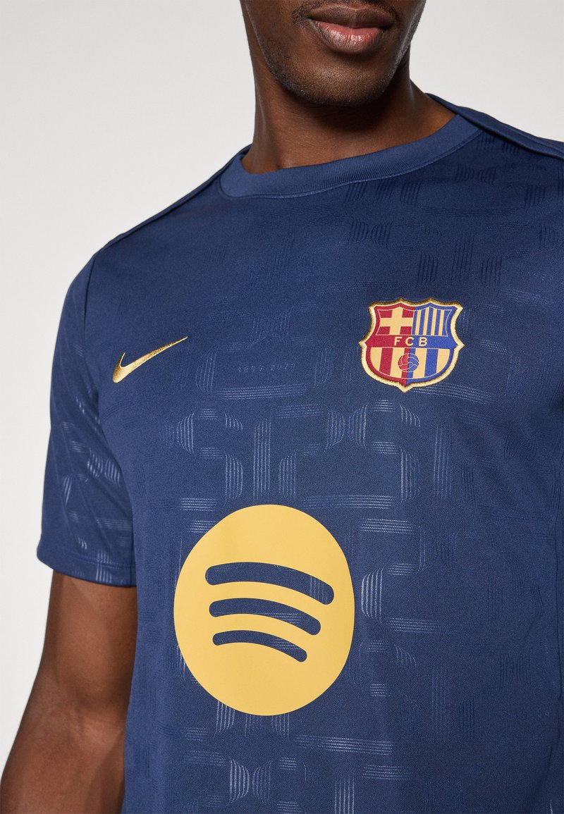 Camisa deportiva azul marino con acentos dorados, que presenta un patrón texturizado, un escudo del club en el pecho y un gran logo amarillo de Spotify.