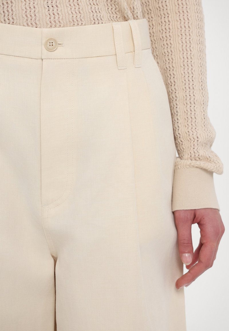 Beżowe spodnie o dopasowanym kroju, z płaskim przodem, kieszeniami bocznymi i pojedynczym zapięciem na guzik w talii. Gładka faktura.