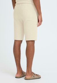 Shorts beige en tricot avec une poche arrière, coupe ajustée et tissu texturé. Portés avec des sandales marron sur un mannequin contre un fond clair.