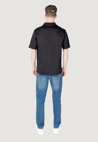 Polo shirt nera corta con colletto, abbinata a jeans in denim azzurro chiaro. Modello visto di spalle, che mostra una vestibilità rilassata e un design di tasca standard.