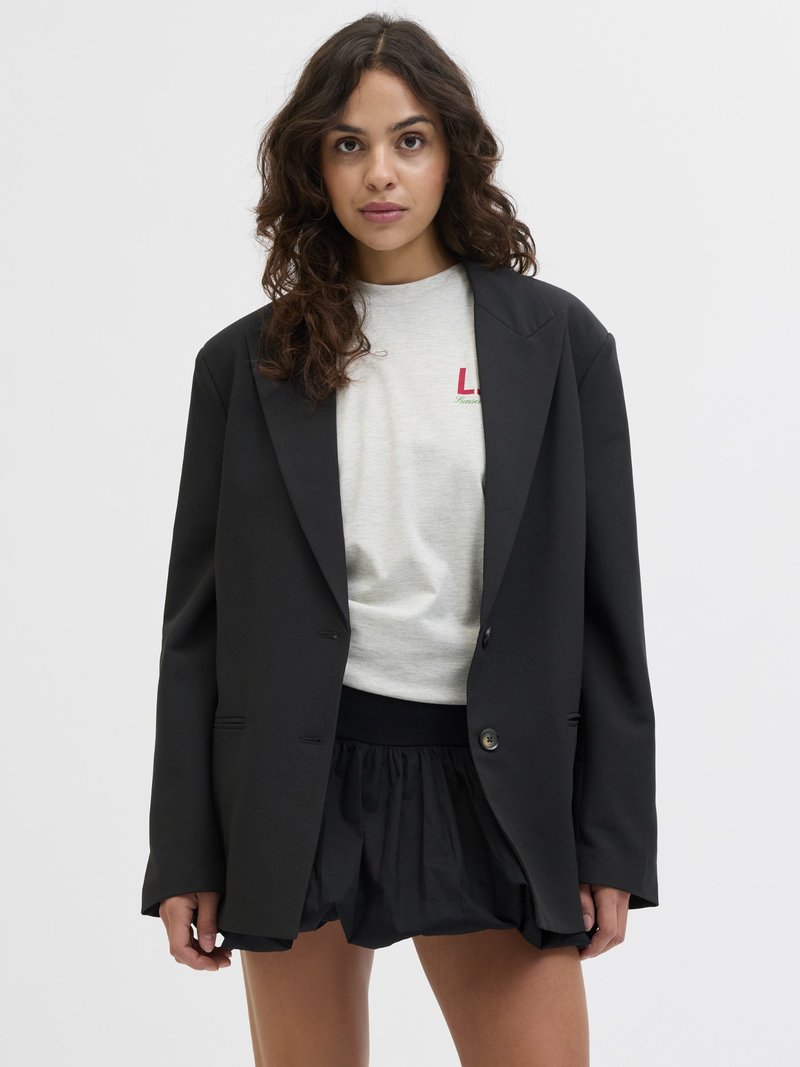 Svart blazer med en strukturerad design, som har två framskjutna knappar och en skräddarsydd passform. Bärs över en ljusgrå sweatshirt och lager-på-lager med en svart kjol.