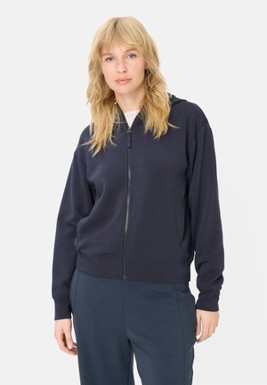 Blonde woman wearing a navy blue zip-up hoodie and matching pants, standing against a plain white background.