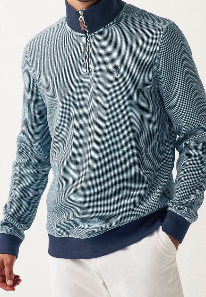 Pull bleu clair en tissu texturé avec col bleu marine. Comprend une demi-fermeture éclair, des poignets et un ourlet côtelés. Logo de la marque discret sur la poitrine.