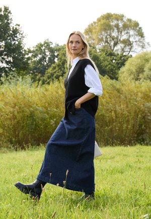 Chaleco de punto negro sobre una camisa blanca, combinado con una falda maxi de mezclilla oscura. Botines negros y un bolso ligero completan el conjunto.