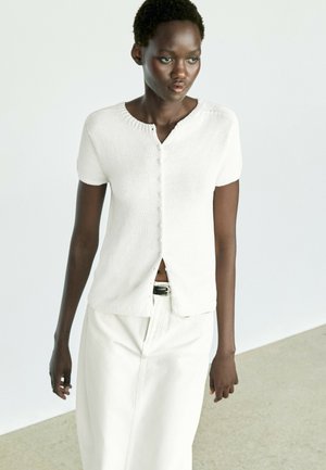 Femme portant un haut blanc à manches courtes boutonné et une jupe blanche avec une ceinture noire, debout sur un fond clair uni.
