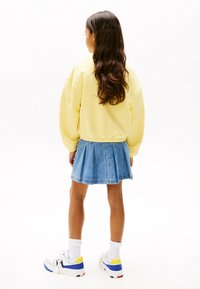 Sudadera amarilla, falda de mezclilla, zapatillas blancas con acentos en azul, amarillo y negro. Tela suave, falda con pliegues, corte relajado.