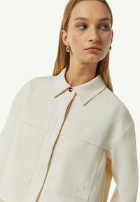 Jasnokremowa kurtka z kołnierzykiem, z przodu na zamek, z dwoma kieszeniami na piersi, subtelną teksturą oraz logo na dolnej części.