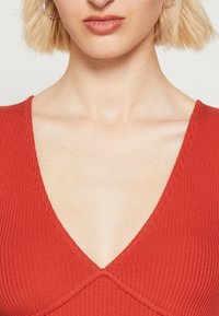 Punainen ribbikangasmekko, jossa on syvä V-muotoinen kaula-aukko ja ristipistoyksityiskohtia. Kangas näyttää teksturoidulta, ja istuva muotoilu korostaa solisluuta.