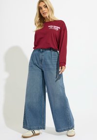 Bordowa koszula z długim rękawem z białym napisem, zestawiona z wysokim stanem, szerokimi niebieskimi dżinsami. Białe sneakersy z czarnymi paskami.