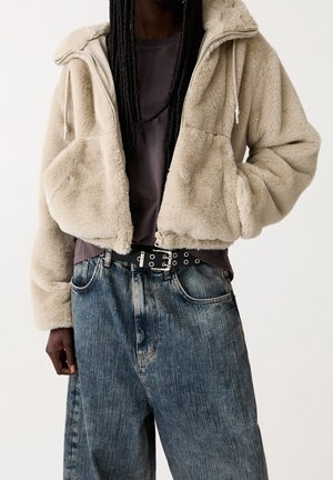 Veste cropped beige, duveteuse, avec une capuche et des poches latérales, portée sur une chemise grise. Associée à un jean bleu délavé taille haute et une ceinture noire.